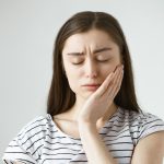 Portrait of unhappy depressed young Caucasian brunette woman closing eyes in frustration and pressing hand against cheek, trying to soothe pain while suffering from toothache, having tooth decay